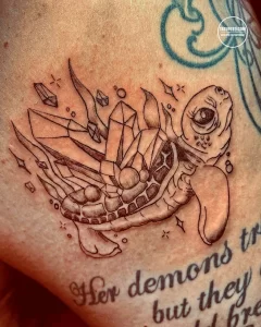 Фото тату на плече: морская черепаха, окруженная кристаллами и надписью "Her demons but they free". Оригинальный рисунок татуировки в черно-белых тонах.