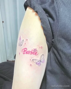 Фото тату с надписью 'Barbie' и двумя розовыми бабочками, украшенными блестящими звездочками, на женском предплечье.