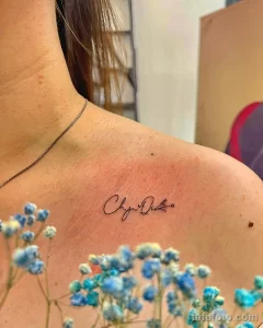 Фото тату: надпись "Chynna Dilia" изящным шрифтом на ключице девушки. Минималистичная татуировка, выполненная в технике лайнуорк.