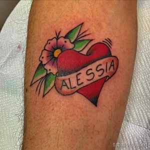 Фотография ноги с татуировкой в виде красного сердца, на котором написано имя "Alessia", окруженного зелеными листьями, демонстрирующая рисунок татуировки в традиционном стиле.