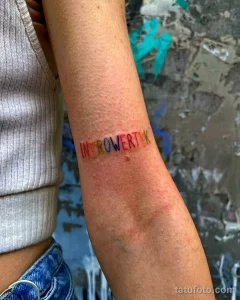 Фотография руки с татуировкой в виде надписи "INTROVERT", буквы которой выполнены в разных ярких цветах, демонстрирующая рисунок татуировки в минималистичном стиле.