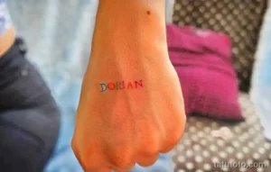 Фотография руки с татуировкой в виде надписи "DORIAN", выполненной цветными чернилами, демонстрирующая рисунок татуировки в минималистичном стиле.