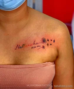 Фото тату с именем "Nattanicha" и датой рождения "28.06.1994" на ключице. Рисунок выполнен в каллиграфическом стиле с добавлением небольших звезд.