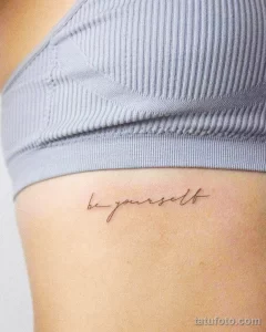 Фото тату: небольшая татуировка надписью 'Be yourself' на боку, выполненная изящным шрифтом черным цветом.
