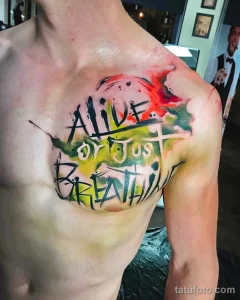 Фото татуировки на груди и плече с надписью 'ALIVE or just BREATHING' выполненной в стиле граффити с использованием ярких красок.