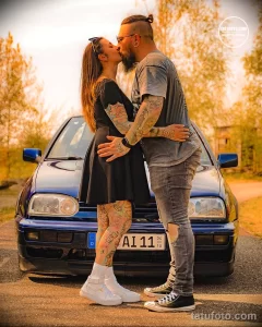 Фотография пары, целующейся на фоне автомобиля, демонстрирующая татуировки на руках. Татуировки выполнены в ярких цветах и изображают различные узоры.
