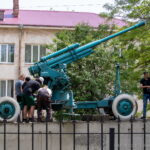 Фото старинной пушки на постаменте. Зеленая окраска, массивные колеса, детализированная конструкция. Исторический памятник на открытом воздухе.