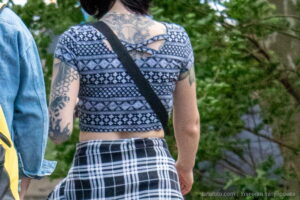 Фото татуировки на спине девушки. Крупный план черно-белого геометрического узора, покрывающего верхнюю часть спины и лопатки. Девушка в клетчатой юбке и топе.