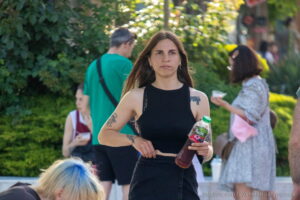 Фотография девушки с татуировкой на плече, идущей по улице. Татуировка выполнена в черном цвете и имеет абстрактный дизайн. Девушка держит в руке бутылку напитка.