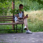 Фотография мужчины, сидящего на скамейке в парке, с татуировкой на руке и смотрящего в смартфон. Зеленый фон листвы.