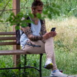 Фотография мужчины, сидящего на скамейке в парке, с татуировкой на руке и смотрящего в смартфон. Солнечный день и зеленая листва.