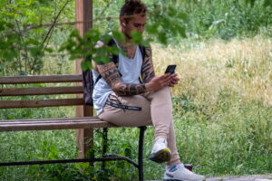 Фотография мужчины, сидящего на скамейке в парке, с татуировкой на руке и смотрящего в смартфон. Солнечный день и зеленая листва.