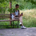 Фотография мужчины, сидящего на скамейке в парке, с татуировкой на руке и смотрящего в смартфон. Зеленый фон листвы.