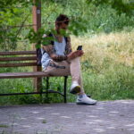 Фотография мужчины, сидящего на деревянной скамейке в парке, с татуировкой на руке и смотрящего в смартфон. Спокойная атмосфера.