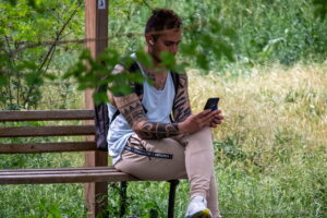 Фотография мужчины, сидящего на скамейке в парке с татуировкой на руке и смотрящего в смартфон. Яркий солнечный день.