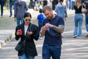 Фотография мужчины с татуировкой на руке и женщины, идущих по улице. Мужчина смотрит в телефон, а женщина держит стакан.
