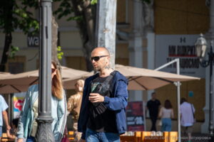 Фото мужчины с татуировкой на шее, одетого в куртку и очки, на фоне городской улицы.