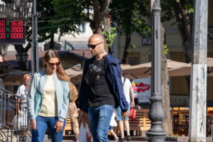 Фото мужчины с татуировкой на шее, одетого в куртку и очки, на фоне городской улицы.
