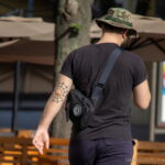 Фото тату паука на плече мужчины, идущего по улице. Татуировка выполнена в черных тонах и выглядит детализированной, с реалистичной прорисовкой паутины.