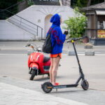 Фотография девушки на электросамокате с татуировкой на ноге, стоящей рядом с красным скутером на городской улице.