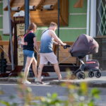 Фотография мужчины, толкающего детскую коляску, с видимой татуировкой на икре ноги. Современный городской пейзаж.