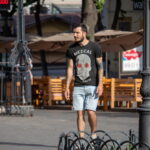 Фото мужчины с татуировкой на руке, идущего по улице. Рука покрыта татуировкой в стиле трайбл, на футболке изображение черепа.