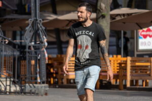 Фото мужчины, идущего по улице в футболке с изображением черепа и с татуировкой на руке в стиле трайбл.
