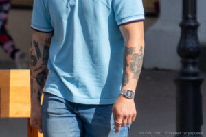 Фото татуировок на руках мужчины в голубой футболке. Видны детализированные рисунки в черном цвете, включая растительные мотивы и изображения животных.