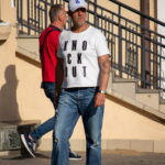 Фото мужчины с татуировкой на руке, одетого в белую футболку, джинсы и бейсболку LA, стоящего рядом с другим мужчиной.