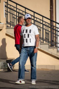 Фото мужчины с татуировкой на руке, одетого в белую футболку, джинсы и бейсболку LA, стоящего рядом с другим мужчиной.