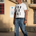 Фото мужчины с татуировкой на руке, одетого в белую футболку, джинсы и кеды. Видна татуировка в стиле олдскул на предплечье.