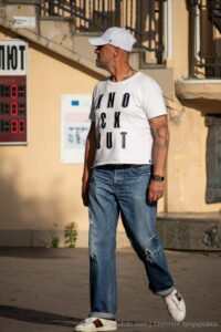 Фото мужчины с татуировкой на руке, одетого в белую футболку, джинсы и кеды. Видна татуировка в стиле олдскул на предплечье.