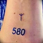 Фото тату на руке: простой черный символ, напоминающий букву 'Y', над числом '580'. Минималистичный рисунок татуировки.