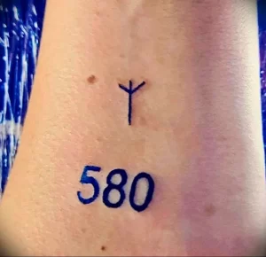 Фото тату на руке: простой черный символ, напоминающий букву 'Y', над числом '580'. Минималистичный рисунок татуировки.