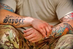 Фотография татуировки на руке мужчины: надпись "Sapper" выполненная красивым шрифтом, и рядом изображение морского пейзажа с кораблем и волнами.