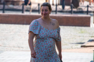 Женщина в цветном платье с открытым плечом, на котором видна небольшая татуировка. Яркий солнечный день, городская среда.