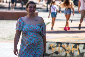 Женщина в цветном платье с открытым плечом, на котором видна небольшая татуировка. Яркий солнечный день, городская среда.