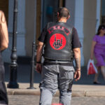 Фотография байкера в кожаной жилетке с нашивкой R.E.D. и татуировкой на руке. Мужчина стоит спиной к камере на городской улице.