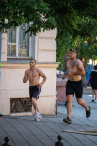 Фотография мужчины, бегущего по улице с обнаженным торсом, на котором видны небольшие татуировки. Он одет в спортивные шорты и наушники.