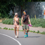 Фотография девушки с татуировкой на руке, идущей по улице с мужчиной. Девушка одета в розовое платье, мужчина несет коврик для йоги.