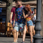 Фотография мужчины с татуировкой на ноге, идущего по улице с девушкой, в яркой рубашке с тропическим принтом.