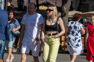 Фотография людей, идущих по улице. У девушки на плече видна небольшая татуировка в виде рисунка. Яркий солнечный день.