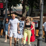 Фотография людей, идущих по улице. У девушки на плече видна небольшая татуировка в виде рисунка. Яркий солнечный день.