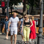 Фотография людей, идущих по улице. У девушки на плече видна небольшая татуировка в виде рисунка. Яркий солнечный день.