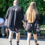 Фотография пары, идущей по улице, держась за руки. Видны татуировки на руках и ногах мужчины, а также татуировка на лодыжке женщины. Солнечный день.
