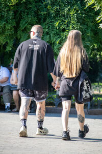 Фотография пары, идущей по улице, держась за руки. Видны татуировки на руках и ногах мужчины, а также татуировка на лодыжке женщины. Солнечный день.