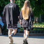 Фотография пары, идущей по парку, демонстрирующая татуировки на руках и ногах мужчины и татуировку на лодыжке женщины. Яркий солнечный день.