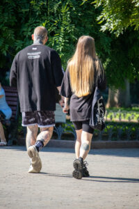 Фотография пары, идущей по парку, демонстрирующая татуировки на руках и ногах мужчины и татуировку на лодыжке женщины. Яркий солнечный день.