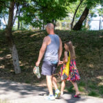 Фото мужчины с татуировкой скорпиона на спине, идущего с девочкой по парку.