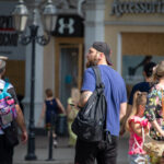 Фото тату на руке мужчины, идущего по улице. Татуировка выполнена в темных тонах, изображает детализированный рисунок. Мужчина одет в кепку и синюю футболку.